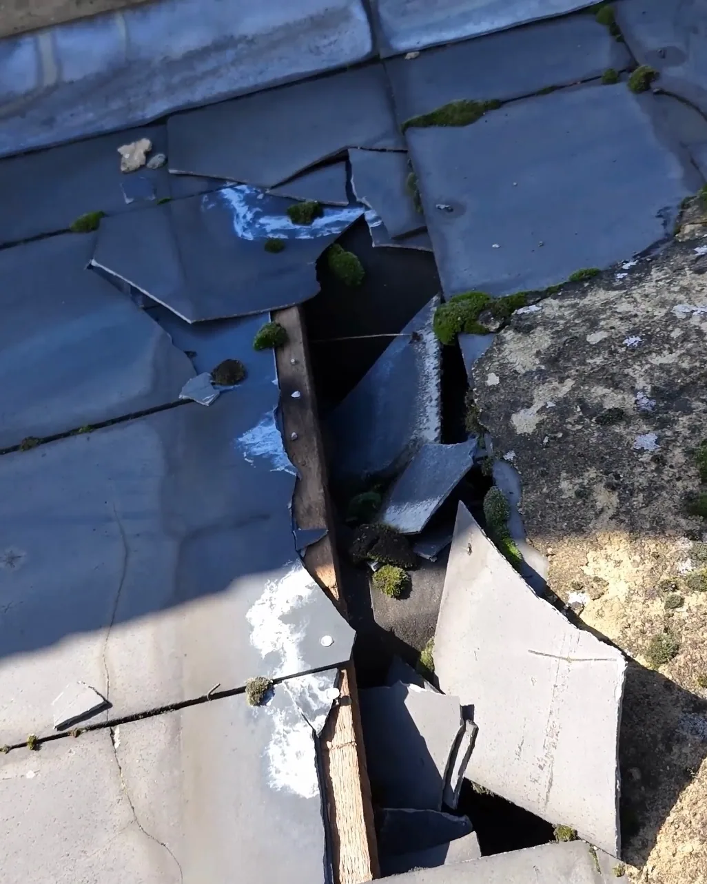 Close drone image of a damaged flat roof, with broken sections of covering and moss along the joints