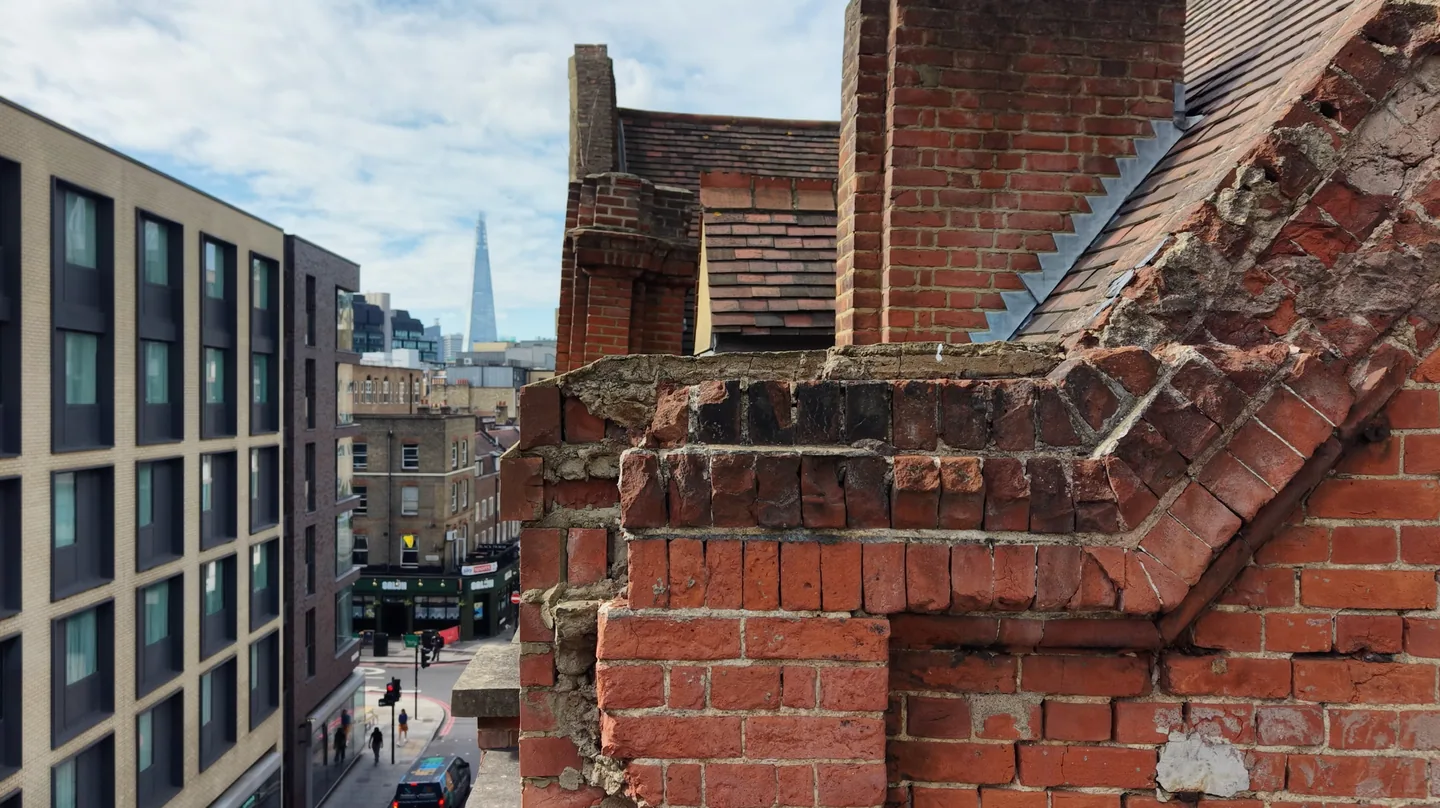 Close drone view of a crumbling red-brick parapet, with eroded mortar, spalled bricks and broken coping, and the Shard in the distance