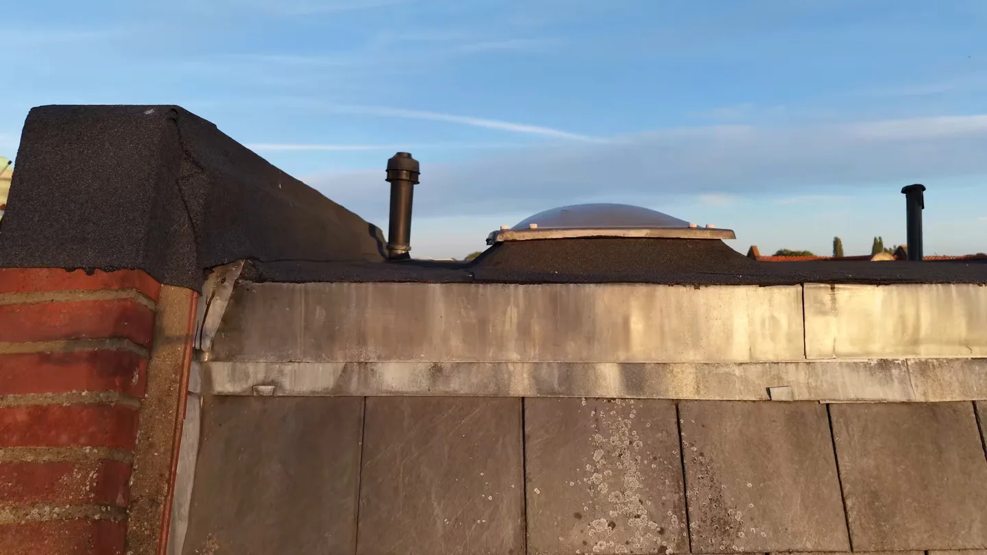 Drone view along a flat roof at sunset, with a weathered parapet coping, a dome rooflight and vent pipes