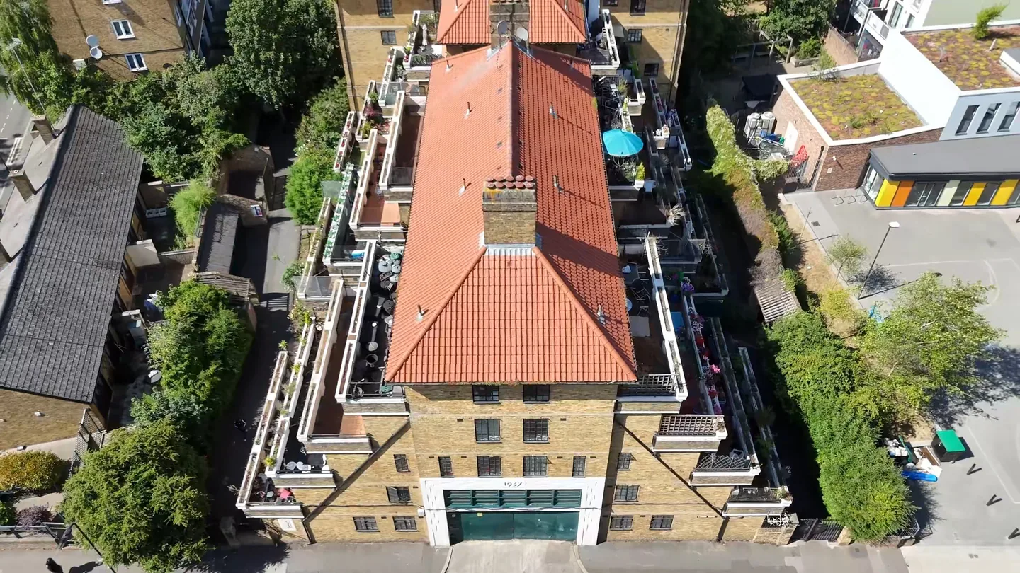 Drone view looking down on a London mansion block, with a red clay-tiled hipped roof, chimney stack and stepped balconies, surrounded by trees