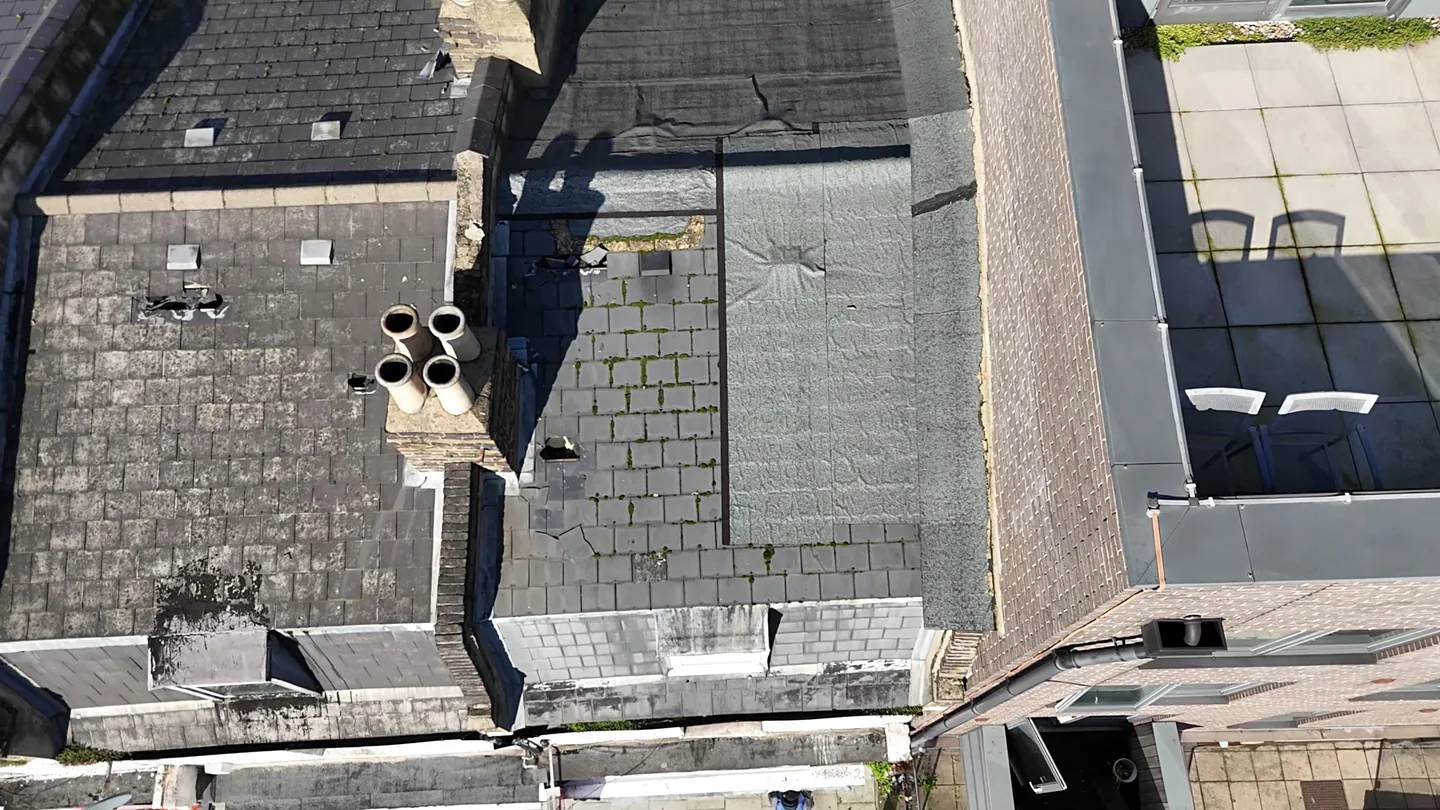 Top-down drone view of London roofs showing slipped and missing slates, a patched felt flat roof with moss, and a chimney stack