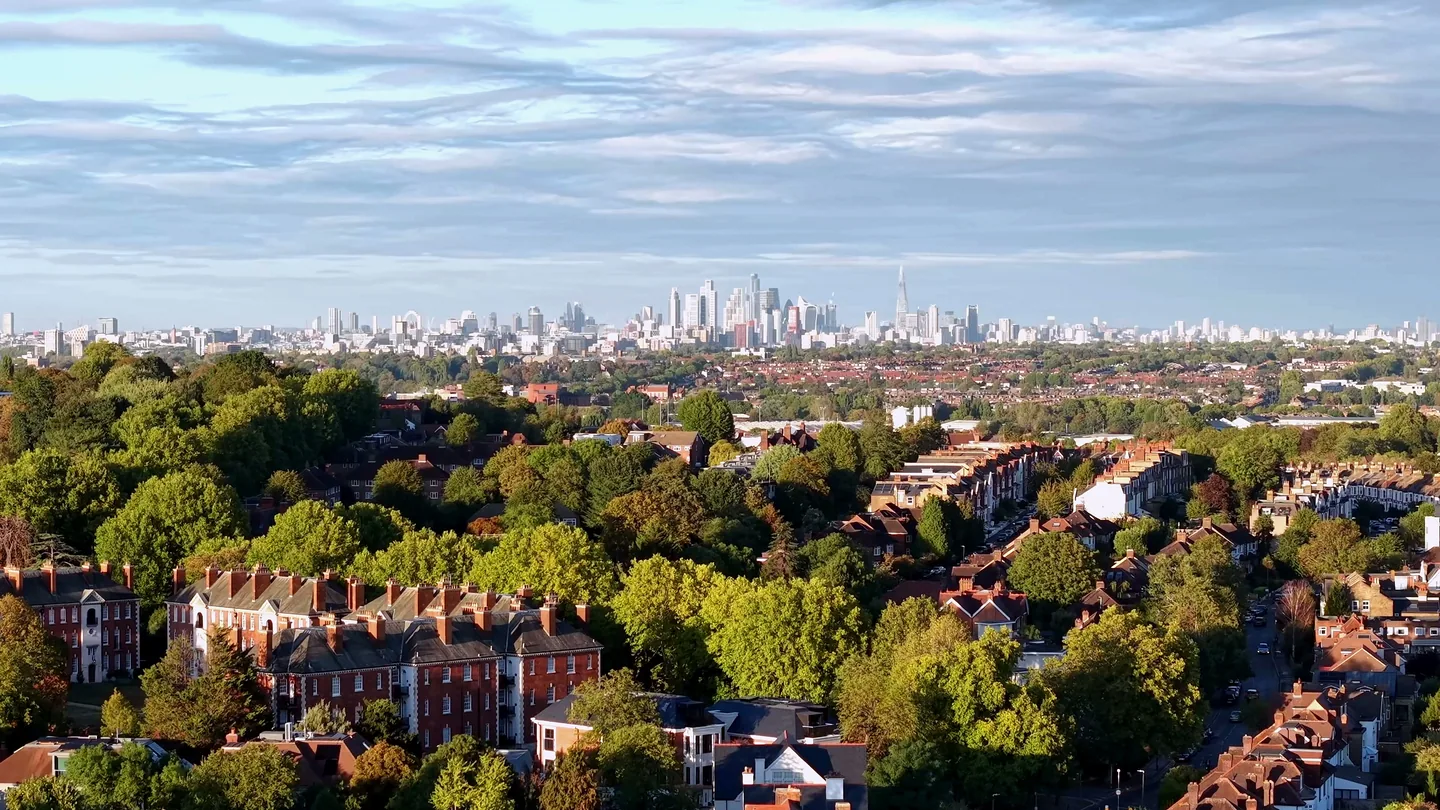 Aerial drone view over tree-lined London streets and terraced housing, with the City of London skyline on the horizon