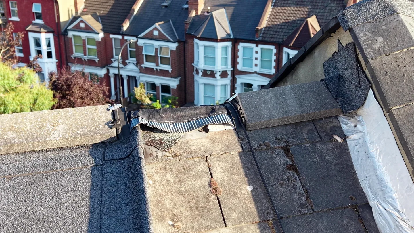 Drone close-up of a torn ridge roll and damaged felt where a flat roof meets the coping, with terraced houses below