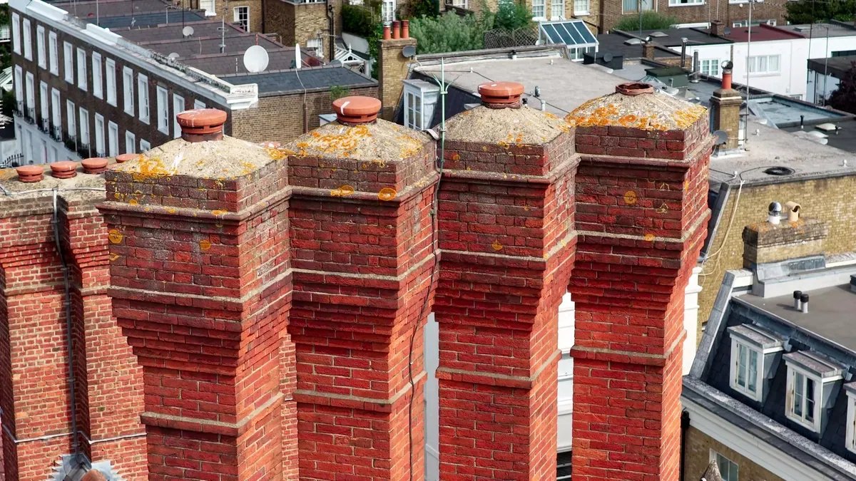 Row of tall red-brick chimney stacks on a London roof, with lichen and weathering on the stone cappings