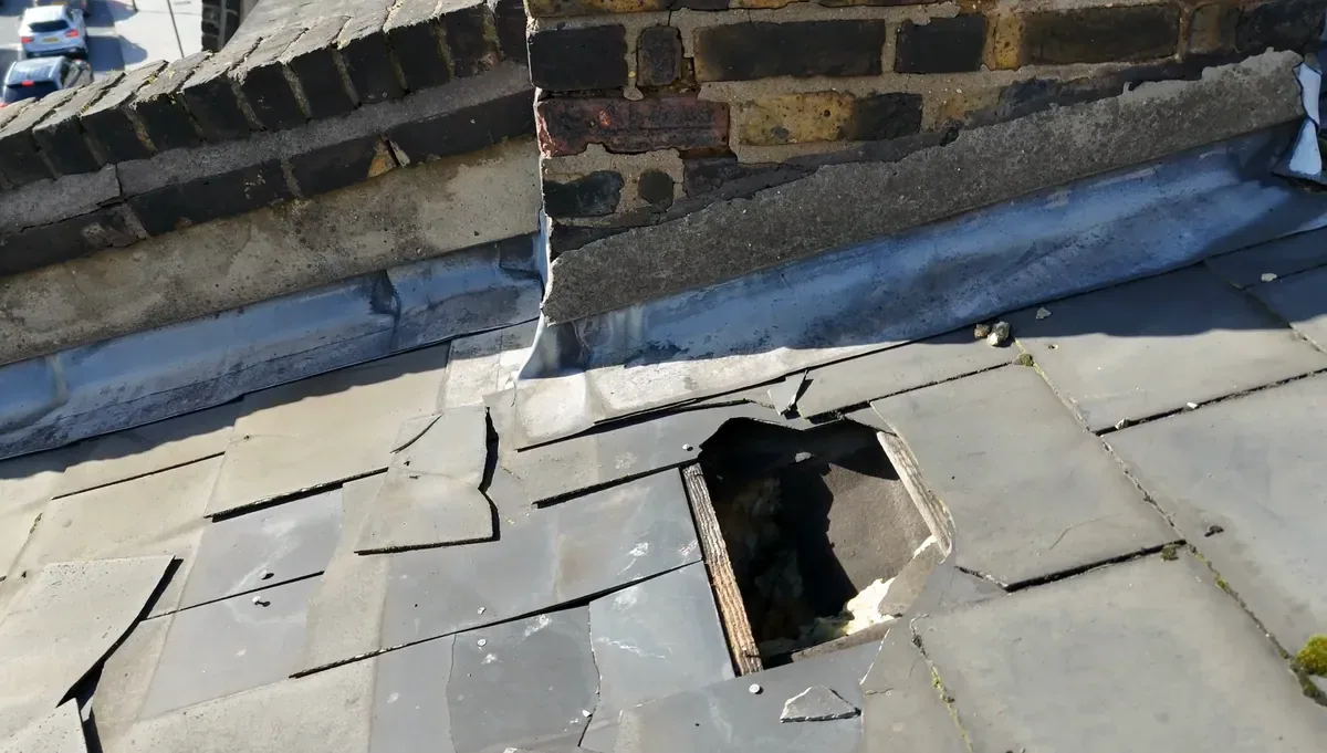 Close drone view of a flat roof where a section of covering has broken away, leaving a hole beside a brick parapet